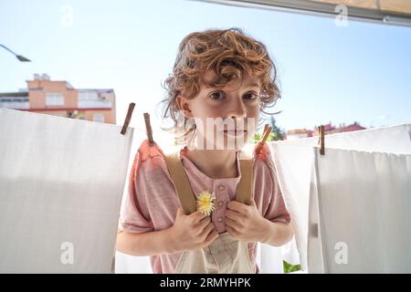 Adorable enfant préadolescent avec fleur jaune s'amusant tout en faisant la lessive et se pendant sur la corde à linge près des draps blancs avec pinces à linge sur bal Banque D'Images