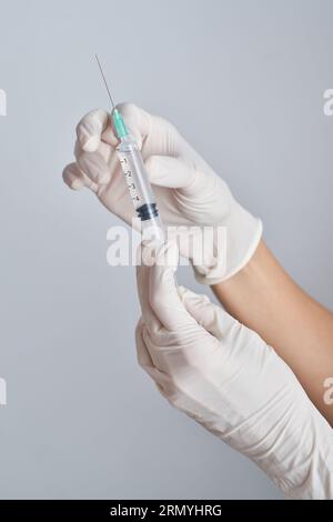 Mains de la culture anonyme médic dans des gants en latex tenant la seringue avec le vaccin sur fond blanc dans la salle lumineuse pendant la pandémie Banque D'Images