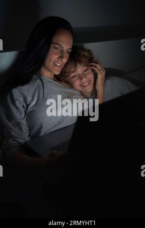 Garçon souriant avec la mère surfant Internet sur netbook tout en faisant ses devoirs ensemble sur le lit dans la chambre sombre tard dans la nuit Banque D'Images