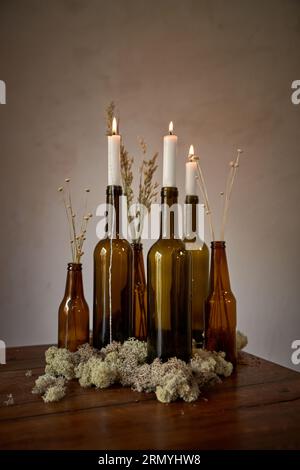 Ensemble de bouteilles en verre vides avec des bougies allumées blanches placées sur la table avec des fleurs séchées près du mur dans la salle lumineuse Banque D'Images