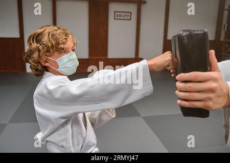 Enfant concentré portant un masque médical de protection et kimono frappant le tampon de frappe pendant l'entraînement de karaté avec entraîneur de culture en dojo Banque D'Images