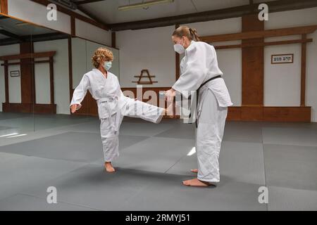 Jeune femme en kimono et masque médical faisant du Krav Maga avec un jeune garçon dans une salle spéciale pour les cours Banque D'Images