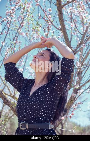 Du dessous de la femme souriante en robe pointillée avec ceinture regardant vers le haut et debout sous l'arbre de lichen avec des branches couvertes de feuilles et rose fleuri Banque D'Images