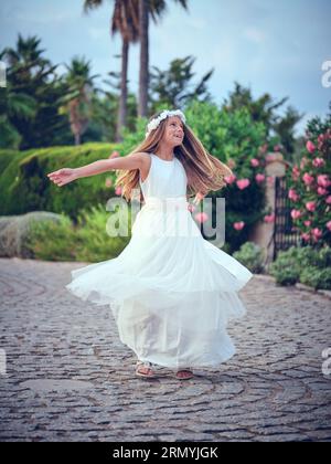 Corps entier de fille préteen positive portant robe blanche élégante et couronne de fleurs tournant autour dans le jardin d'été et regardant loin Banque D'Images