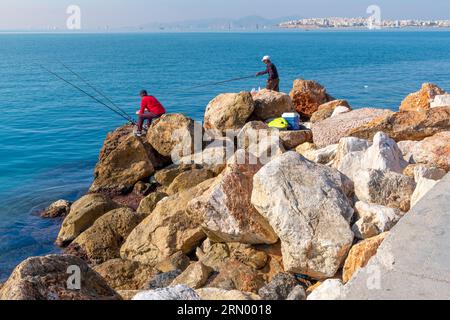 Deux hommes pêchent au large des rochers de bord de mer et un chat se fond dans les rochers en contrebas dans la région de la Riviera athénienne, Paleo Faliro, près d'Athènes, Grèce. Banque D'Images