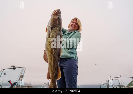 Heureuse pêcheuse tenant une grosse morue arctique. Norvège pêche heureuse. Femme avec du poisson de morue dans les mains Banque D'Images