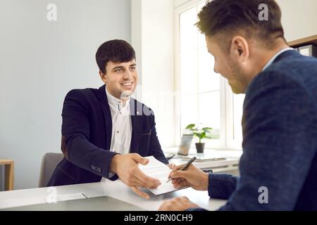 Manager souriant positif donnant le contrat à signer à son client lors d'une réunion d'affaires dans le bureau. Banque D'Images