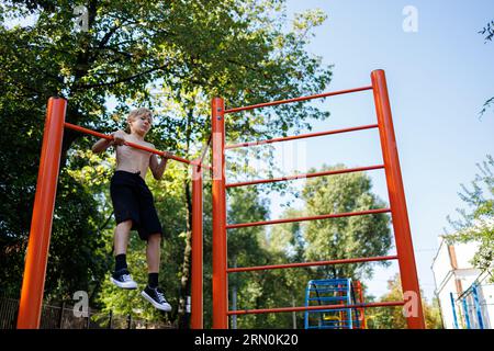 Le garçon de sport effectue des tractions sur la barre horizontale. Séance d'entraînement de rue sur une barre horizontale dans le parc de l'école. Banque D'Images