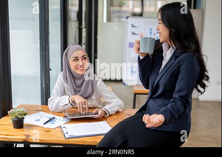 An attractive and smiling Asian Muslim female office worker wearing a hijab enjoys chatting with her colleague during a coffee break in the office. Banque D'Images