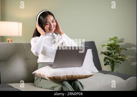 Une belle femme asiatique portant des écouteurs parle lors d'un appel vidéo avec son ami à travers son ordinateur portable sur un canapé dans le salon. réunion en ligne, Banque D'Images