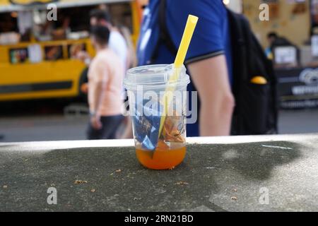 Verre en plastique avec boisson orange, paille et petits morceaux d'emballages de chocolat couchés sur la table comme ordures. Banque D'Images