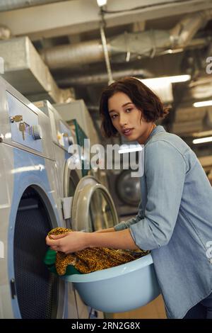 jeune femme asiatique regardant la caméra et tenant le panier avec des vêtements dans la blanchisserie publique Banque D'Images