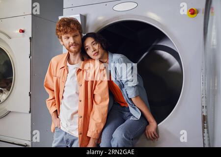 jeune femme asiatique assise sur la machine à laver près du petit ami rousse dans la blanchisserie publique Banque D'Images