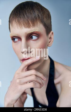 modèle queer avec maquillage regardant loin sur fond gris, personne androgyne, portrait, identité Banque D'Images