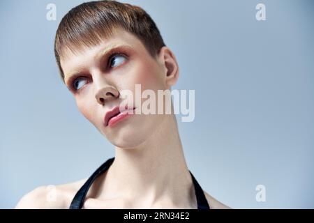 portrait, modèle androgyne avec maquillage regardant loin sur fond gris, beauté et visage, queer Banque D'Images