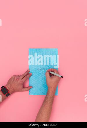 Vue aérienne des mains masculines adultes caucasiennes écrivant le mot Awesome sur une enveloppe d'emballage à bulles bleues. Fond rose avec de la place pour l'espace de copie. Banque D'Images