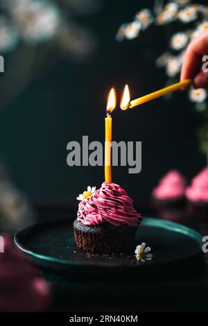 Cupcake au chocolat avec glaçage rose décoré de fleurs comestibles. Main recadrée allumant une bougie avec une autre bougie sur le dessus du cupcake. Banque D'Images