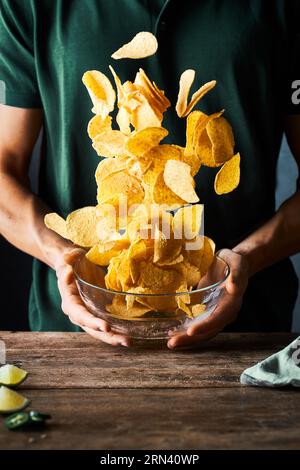 Gros plan d'une personne tenant un bol de croustilles tortilla les jetant en l'air. Des mouvements figés captivants. Il y a une table en bois rustique. Banque D'Images