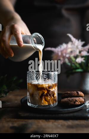 Main coupée en pousant le lait de la bouteille en verre dans un verre de café faisant du café glacé. C'est sur une vieille table en bois rustique. Banque D'Images