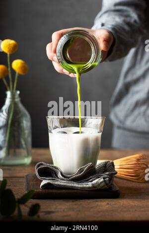 Main coupée versant une dose de matcha dans un verre de lait pour préparer du matcha latte. Table en bois et fond gris. Salle de séjour ou salle à manger Banque D'Images
