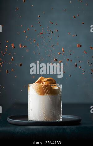 Café Dalgona dans un verre moderne texturé sur un fond gris foncé entouré de poudre de café instantané tombant (explosant) et de ses morceaux. Banque D'Images