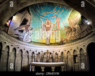 WASHINGTON, DC, États-Unis — Une mosaïque détaillée représentant la Résurrection de Jésus-Christ orne le plafond en demi-dôme au-dessus de l'autel dans la chapelle de la Résurrection de la cathédrale nationale de Washington, DC. La chapelle, construite dans le style normand, présente des espaces intimes avec des arches semi-circulaires et des plafonds inférieurs. Cette mosaïque, créée par l'artiste Hildreth Meirère, fait partie d'une série illustrant les apparitions post-Résurrection de Jésus, avec des mosaïques supplémentaires conçues par Rowan et Irene LeCompte sur les parois latérales. Banque D'Images