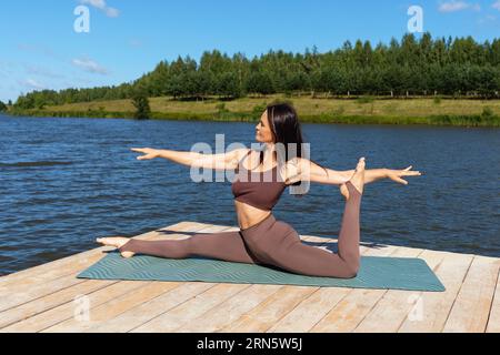 Une femme pratiquant le yoga, effectue une variation de l'exercice Hanumanasana, une division longitudinale, s'entraîne dans des vêtements de sport séparés tout en étant assise sur un woo Banque D'Images