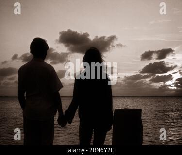 1970s BACK VIEW OF ANONYMOUS SILHOUETTED TEEN COUPLE HOLDING HANDS LOOKING OUT TO SUNSET OVER WATER - r22264 HAR001 HARS FEMALES RURAL COPY SPACE FRIENDSHIP HALF-LENGTH PERSONS CARING MALES TEENAGE GIRL TEENAGE BOY SILHOUETTES B&W OUTLINE DREAMS SILHOUETTED HOLDING HANDS REAR VIEW RELATIONSHIPS CONNECTION CONCEPTUAL FROM BEHIND TEENAGED ANONYMOUS PERSONAL ATTACHMENT AFFECTION BACK VIEW EMOTION JUVENILES TOGETHERNESS BLACK AND WHITE HAR001 OLD FASHIONED Stock Photo