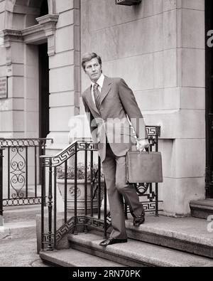 HOMME D'AFFAIRES DES ANNÉES 1970 PORTANT UN COSTUME ET UNE CRAVATE TENANT UNE MALLETTE MARCHANT VERS LE BAS DES MARCHES DU BÂTIMENT AVEC UNE RAMPE EN FER FORGÉ À LA RECHERCHE D'UNE CABINE - S20461 HAR001 HARS PROFESSION CONFIANCE B&W COSTUME ET CRAVATE OCCUPATION VENTE DE STYLES ET CARRIÈRES OCCUPATIONS EXTÉRIEURES DE GARDE-CORPS IMMOBILIER ÉLÉGANT AVOCAT MODE PRUDENTE MI-ADULTE MI-ADULTE HOMME VENDEURS FORGÉ NOIR ET BLANC CAUCASIEN SORTANT DE HAR001 OLD FASHIONED UNSOURIANT Banque D'Images