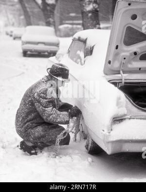 1960s MAN PUTTING CHAINS ON CAR TIRES DURING SNOW STORM - m6872 HAR001 HARS EXTERIOR CHAINS AUTOMOBILES VEHICLES WINTERY TIRES BLACK AND WHITE CAUCASIAN ETHNICITY HAR001 OLD FASHIONED Stock Photo