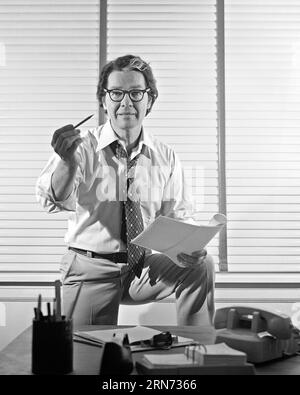 1970s BUSINESSMAN AT DESK WEARING EYEGLASSES NECKTIE LOOSENED SHIRT SLEEVES ROLLED UP HOLDS PENCIL IN ONE HAND PAPERS IN OTHER  - o4155 HAR001 HARS ROLLED STUDIO SHOT MANAGER COPY SPACE HALF-LENGTH PERSONS THOUGHTFUL MALES CONFIDENCE EXECUTIVES EYEGLASSES GESTURING B&W EYE CONTACT STRATEGY EXCITEMENT LEADERSHIP DIRECTION UP GESTURES NECKTIE OCCUPATIONS BOSSES CONCEPTUAL SLEEVES SINCERE LOOSENED SOLEMN FOCUSED GRAY STREAK HOLDS INTENSE MANAGERS MID-ADULT MID-ADULT MAN SOLUTIONS BLACK AND WHITE CAREFUL CAUCASIAN ETHNICITY EARNEST HAR001 INTENT OLD FASHIONED Stock Photo