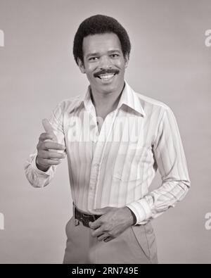 1970s AFRICAN-AMERICAN MAN WITH MUSTACHE SMILING GIVING THUMBS UP GESTURE LOOKING AT CAMERA HAND EXTENDED IN GREETING - s20396 HAR001 HARS THUMBS EYE CONTACT MUSTACHE HAPPINESS AGREE HELLO MUSTACHES AFRICAN-AMERICANS AFRICAN-AMERICAN NETWORKING OKAY BLACK ETHNICITY OPPORTUNITY UP FACIAL HAIR GESTURES APPROVAL FRIENDLY STYLISH THUMBS UP YOUNG ADULT MAN BLACK AND WHITE HAR001 OLD FASHIONED AFRICAN AMERICANS Stock Photo