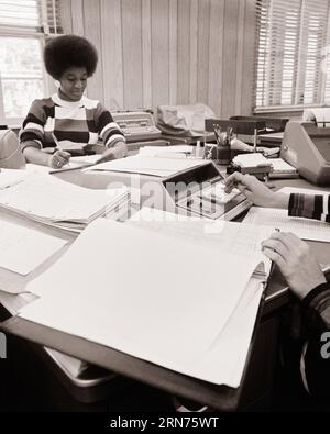 1970s AFRICAN AMERICAN WOMAN WITH AFRO HAIRSTYLE WORKING IN AN ACCOUNTING OFFICE CAUCASIAN HANDS USING AN ELECTRIC CALCULATOR  - o4052 HAR001 HARS B&W GOALS AFRICAN-AMERICANS AFRICAN-AMERICAN HAIRSTYLE LOW ANGLE ADDING MACHINE CLERKS BOOKKEEPER BLACK ETHNICITY OFFICE WORKER OCCUPATIONS USING GAL FRIDAY ACCOUNTING ADMINISTRATOR CALCULATORS SECRETARIES SUPPORT AMANUENSIS BOOKKEEPING COOPERATION MID-ADULT MID-ADULT WOMAN TOGETHERNESS BLACK AND WHITE CAUCASIAN ETHNICITY CLERICAL HANDS ONLY HAR001 OLD FASHIONED AFRICAN AMERICANS Stock Photo