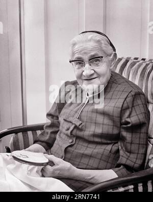 1960s ELDERLY WOMAN WEARING GLASSES SITTING IN CHAIR HOLDING EMBROIDERY IN HOOP SMILING   - s16393 HAR001 HARS EYE CONTACT SENIOR WOMAN EMBROIDERY SKILL ACTIVITY AMUSEMENT HAPPINESS OLD AGE OLDSTERS HIGH ANGLE OLDSTER HOBBY INTEREST AGING HOBBIES KNOWLEDGE PASTIME PRIDE PLEASURE ELDERS CONNECTION GERIATRIC CREATIVITY ELDERLY WOMAN EMBROIDER RELAXATION AMATEUR BLACK AND WHITE CAUCASIAN ETHNICITY ENJOYMENT HAR001 OLD FASHIONED Stock Photo