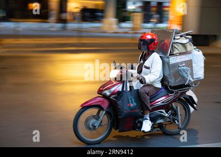 Chauffeur de livraison en mouvement sur la rue de Bangkok à bord d'une moto surchargée de paquets et de sacs la nuit Banque D'Images