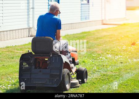 Le travailleur tond le territoire de l'installation industrielle. L'homme conduit la tondeuse à gazon automotrice sur la pelouse le jour ensoleillé d'été. Les clair de lune des retraités âgés comme jardinier. Banque D'Images