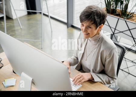 heureuse femme d'affaires mature tapant sur le clavier tout en travaillant sur l'ordinateur dans le bureau, vue à angle élevé Banque D'Images