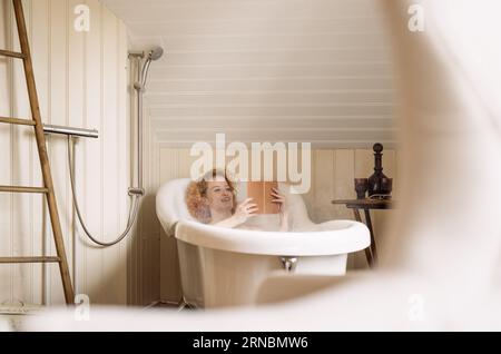 Jeune femme allongée dans la baignoire et lisant le livre à la lumière du jour Banque D'Images