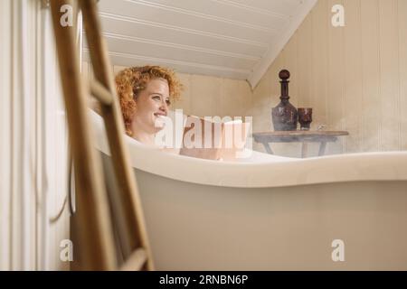 Cheerful young woman lying in bathtub and reading book in daylight Banque D'Images