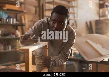 Portrait vue de face d'un jeune homme noir ponçant des meubles en bois dans un atelier de menuiserie, espace copie Banque D'Images