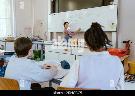 Enseignante souriante écrivant sur le tableau blanc tout en enseignant aux étudiants masculins assis dans la salle de classe Banque D'Images