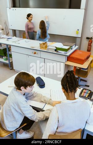 Vue à angle élevé du garçon aidant l'ami masculin tout en étant assis sur le bureau dans la salle de classe Banque D'Images