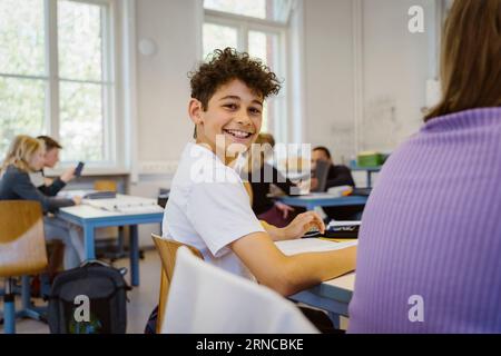 Portrait vue de côté d'écolier souriant assis au bureau dans la salle de classe Banque D'Images