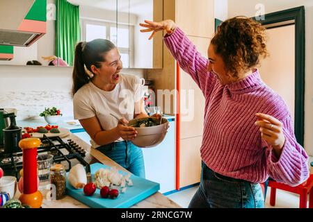 Jeunes amies joueuses appréciant tout en faisant de la salade dans la cuisine à la maison Banque D'Images