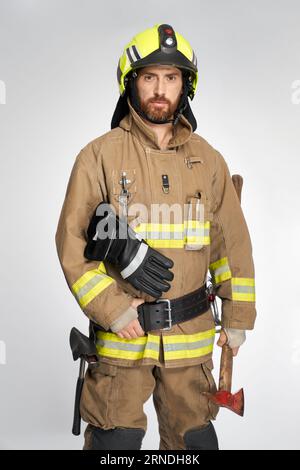 Pompier caucasien attrayant en uniforme posant avec hache rouge en studio. Portrait de pompier barbu confiant avec la main sur la ceinture, regardant la caméra, sur fond gris. Concept de travail, profession. Banque D'Images