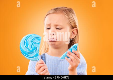 Des enfants, des bonbons et une fille triste avec une sucette cassée sur un fond orange dans le studio semblant bouleversé. Enfants, bonbons et malheureux avec une fille Banque D'Images