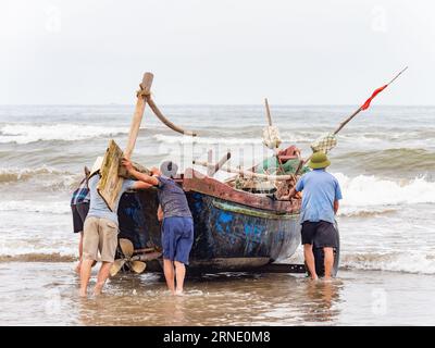 Quatre hommes lancent un bateau de pêche contre le vent et les hautes vagues à Sam son Beach, Thanh Hoa, Vietnam Banque D'Images