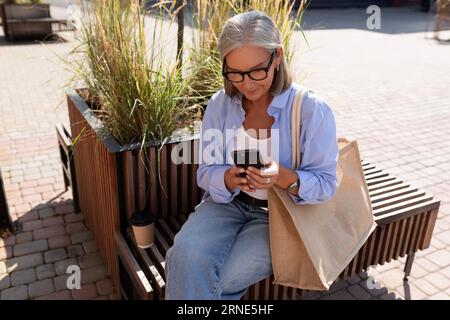 une belle vieille femme mature aux cheveux gris habillée avec élégance s'assoit sur un banc après avoir fait du shopping Banque D'Images