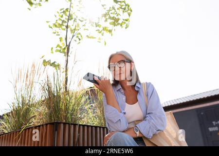 femme mûre aux cheveux gris en tenue d'été s'assoit sur un banc et attend un ami dans la rue Banque D'Images