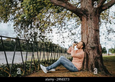 jeune homme assis sur le sol penchez-vous à un arbre et prendre des photos avec son vieil appareil photo rétro en main devant son visage regarder à travers le viseur et profiter du sc Banque D'Images